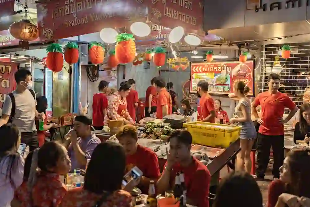 Crowded Asian street food market at night with vendors in red shirts serving customers at outdoor tables, decorated with orange lanterns hanging overhead