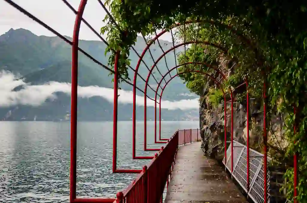 Red metal archway walkway along lakefront with mountains and clouds in background, stone wall and green foliage on right side