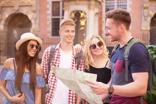 A group of four smiling young tourists standing outside a historic building, holding and looking at an open paper map