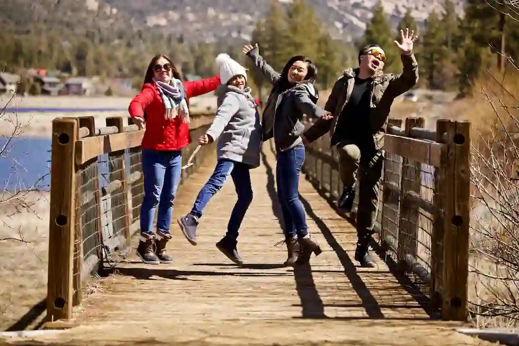 Four friends jumping excitedly on wooden bridge with mountain landscape and pine trees in background