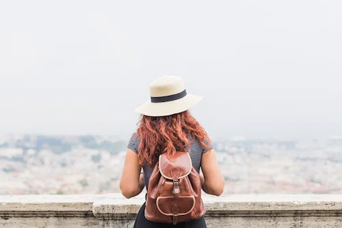 A woman from behind wearing a straw hat and a neatly packed brown leather backpack, looking out over a hazy city from a tower
