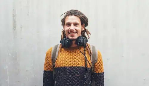A smiling man with dreadlocks, wearing a textured sweater and headphones, stands against a grey wall.