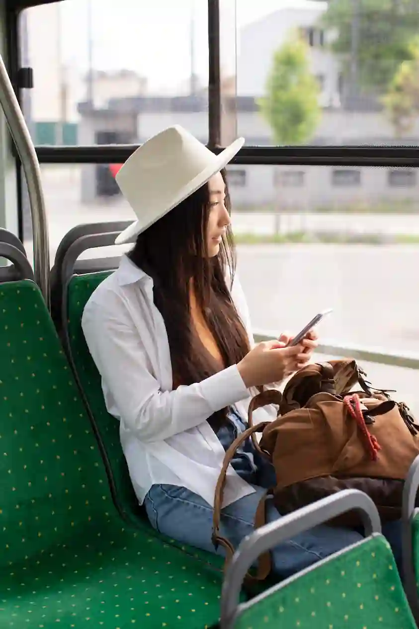 Young woman wearing white hat sitting on public bus checking her phone with travel bag beside her