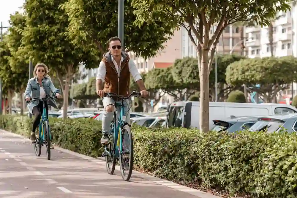 Man and woman cycling on dedicated bike lane through city street lined with green trees and hedges, with vehicles parked along the road
